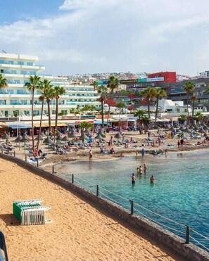 Ein Strand mit klarem, blauem Wasser, Palmen und einer Reihe von Hotels im Hintergrund.
