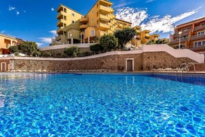 A swimming pool with clear blue water, surrounded by a stone wall and a building with multiple balconies.