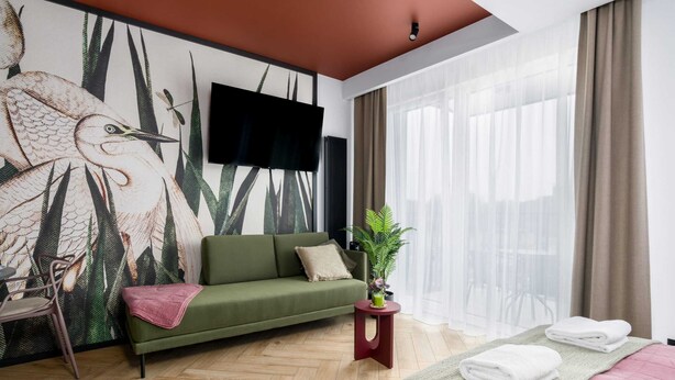 Una habitación de hotel moderna con un sofá verde, un televisor de pantalla plana, una planta en maceta y una cama con toallas dobladas.