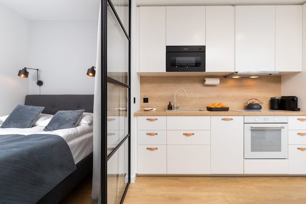 A modern bedroom with a bed, bedside table, and a nightstand. Adjacent to the bedroom is a compact kitchenette with a stove, sink, and countertop.