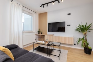 A modern living room with a glass coffee table, a sofa, a TV mounted on the wall, and a potted plant.