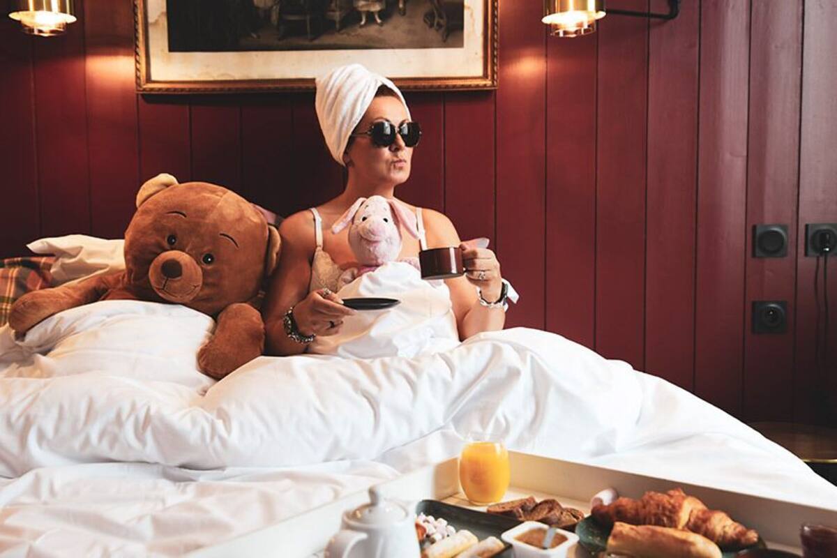 Eine Person sitzt auf einem Bett, neben sich ein Teddybär und eine Tasse, mit Frühstücksleckereien am Rand.