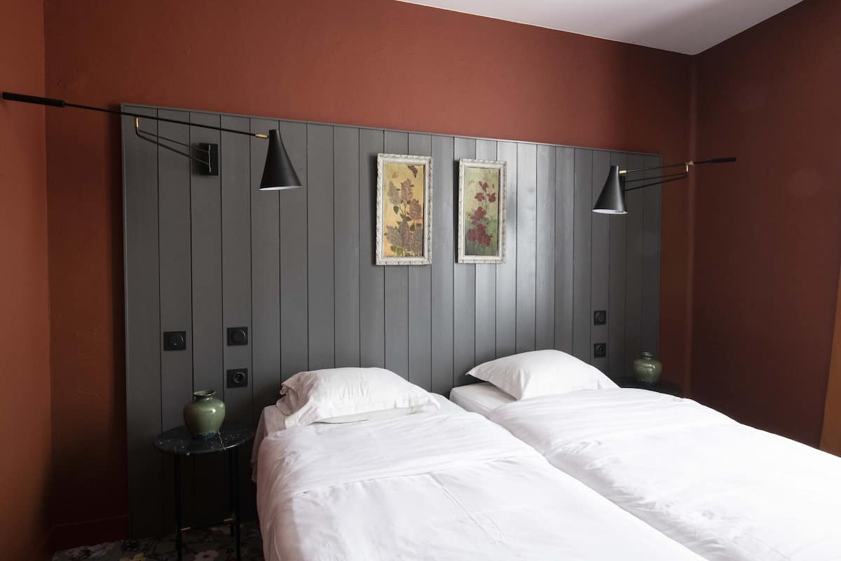 Ein Hotelzimmer mit zwei Betten, einem grauen Kopfteil, zwei Wandleuchten und zwei gerahmten Blumenbildern.