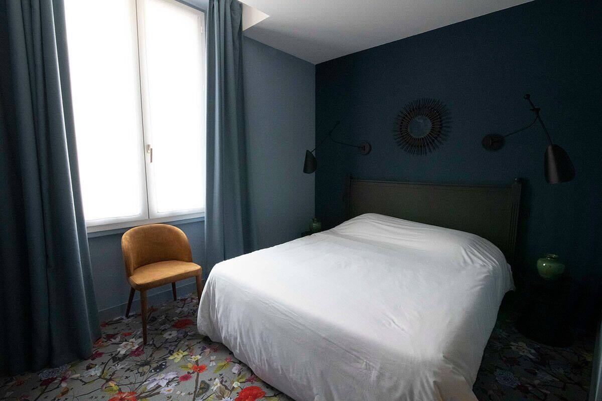 Ein Schlafzimmer mit Bett, Stuhl, einem Fenster mit Vorhängen und einer Wandlampe.