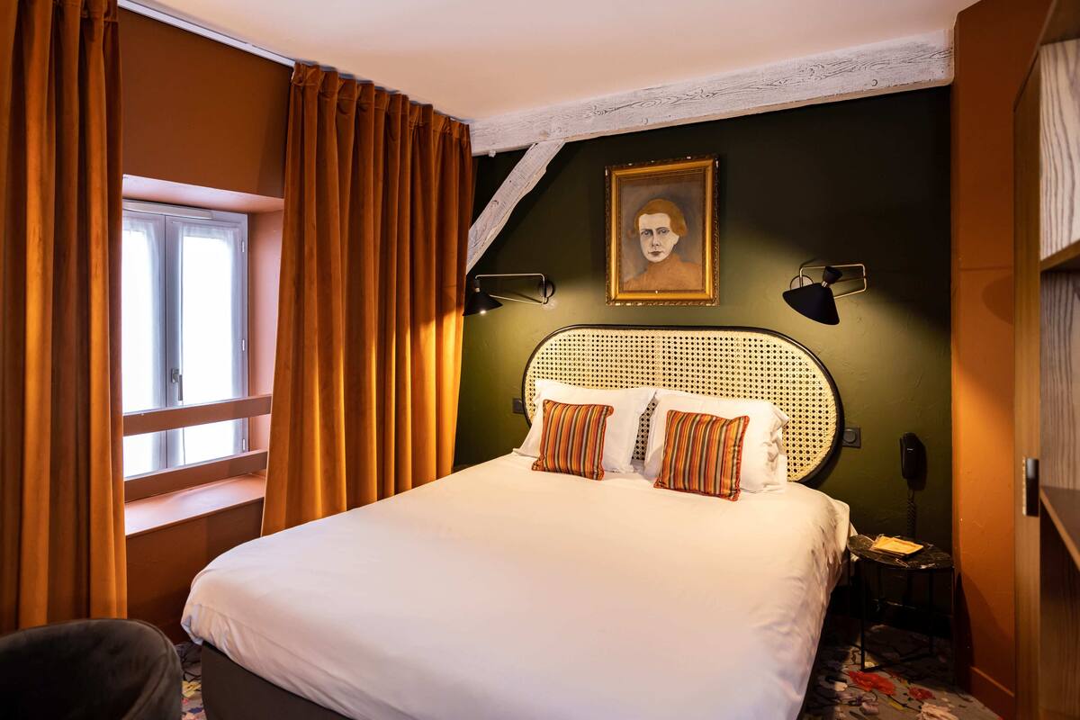 Ein Hotelzimmer mit Bett, Kopfteil, Nachttisch, Lampe, Telefon und einem Fenster mit Vorhängen.