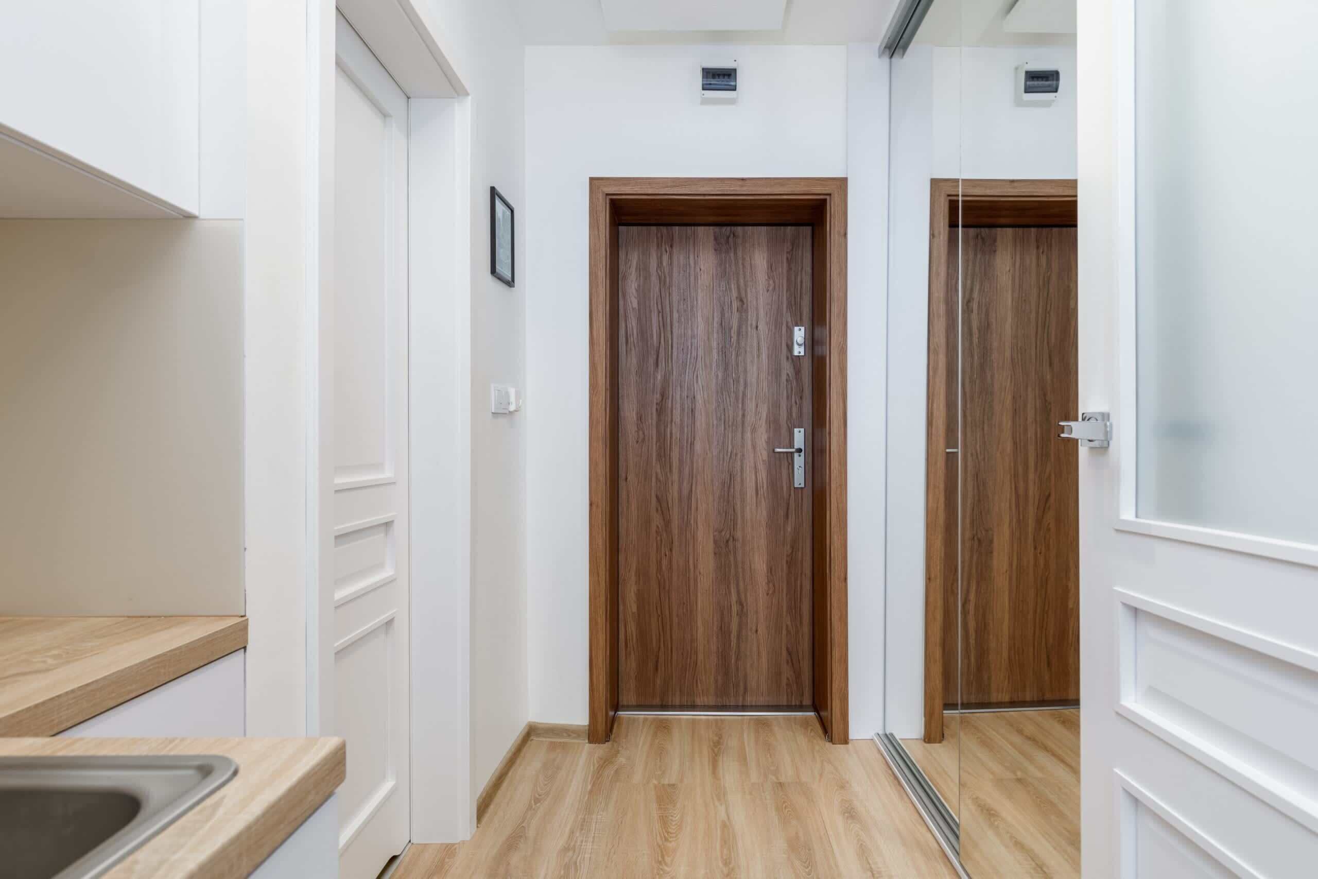 Un couloir étroit avec des portes en bois, un plancher de couleur claire et un coin cuisine équipé d’un évier.