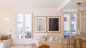 Un soggiorno moderno con un divano, un piccolo sgabello e una sedia di legno. Sono presenti due ampie finestre con vista sugli edifici.
