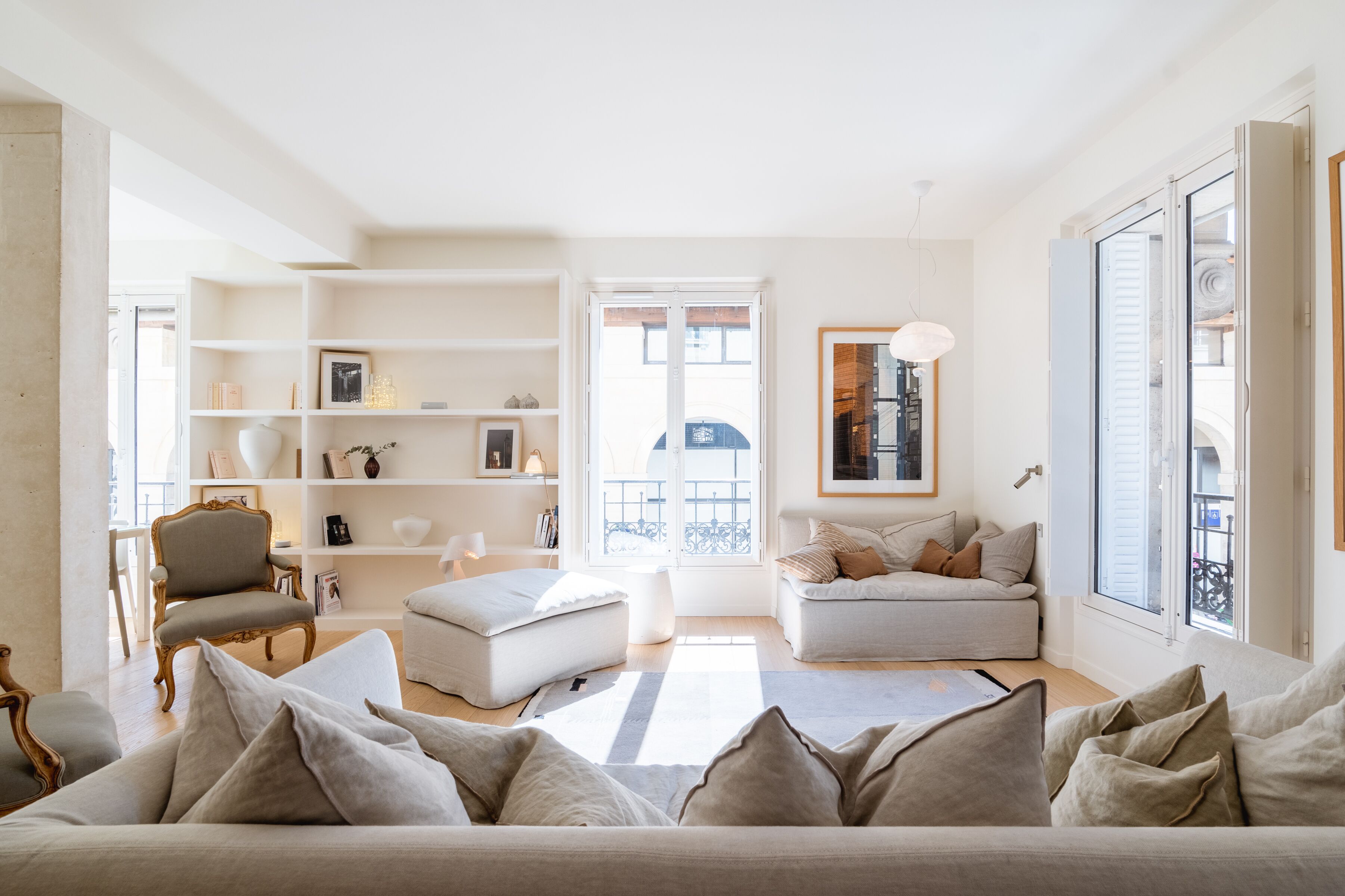 A spacious living room with a large sofa, a smaller sofa, a chair, and a bookshelf. There is a large window with a view of the outside.