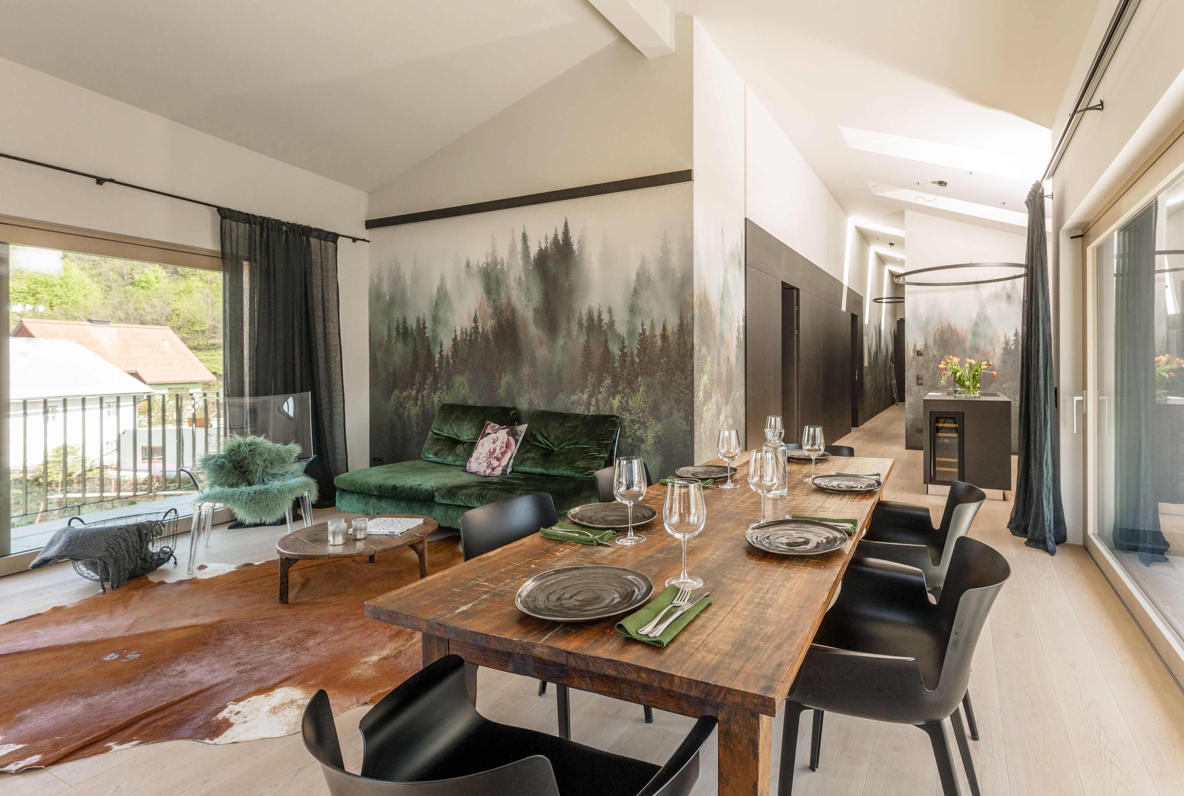 A modern dining area with a wooden table set for six, a green sofa, and a forest mural on the wall.