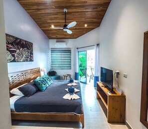A modern bedroom with a bed, a ceiling fan, a TV, and a view of greenery outside.