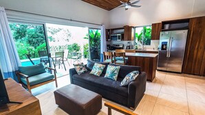 A modern living room with a sofa, ottoman, and a dining area with chairs. A kitchen with a refrigerator and cabinets is visible in the background. - 3 bedroom. 10 min to Beach. Private Pool. (Ojochal)