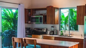 A modern kitchen with wooden cabinets, a microwave, a stove, a coffee maker, and a blender. There is a dining table with chairs and a view of lush greenery through the windows. - 3 bedroom. 10 min to Beach. Private Pool. (Ojochal)