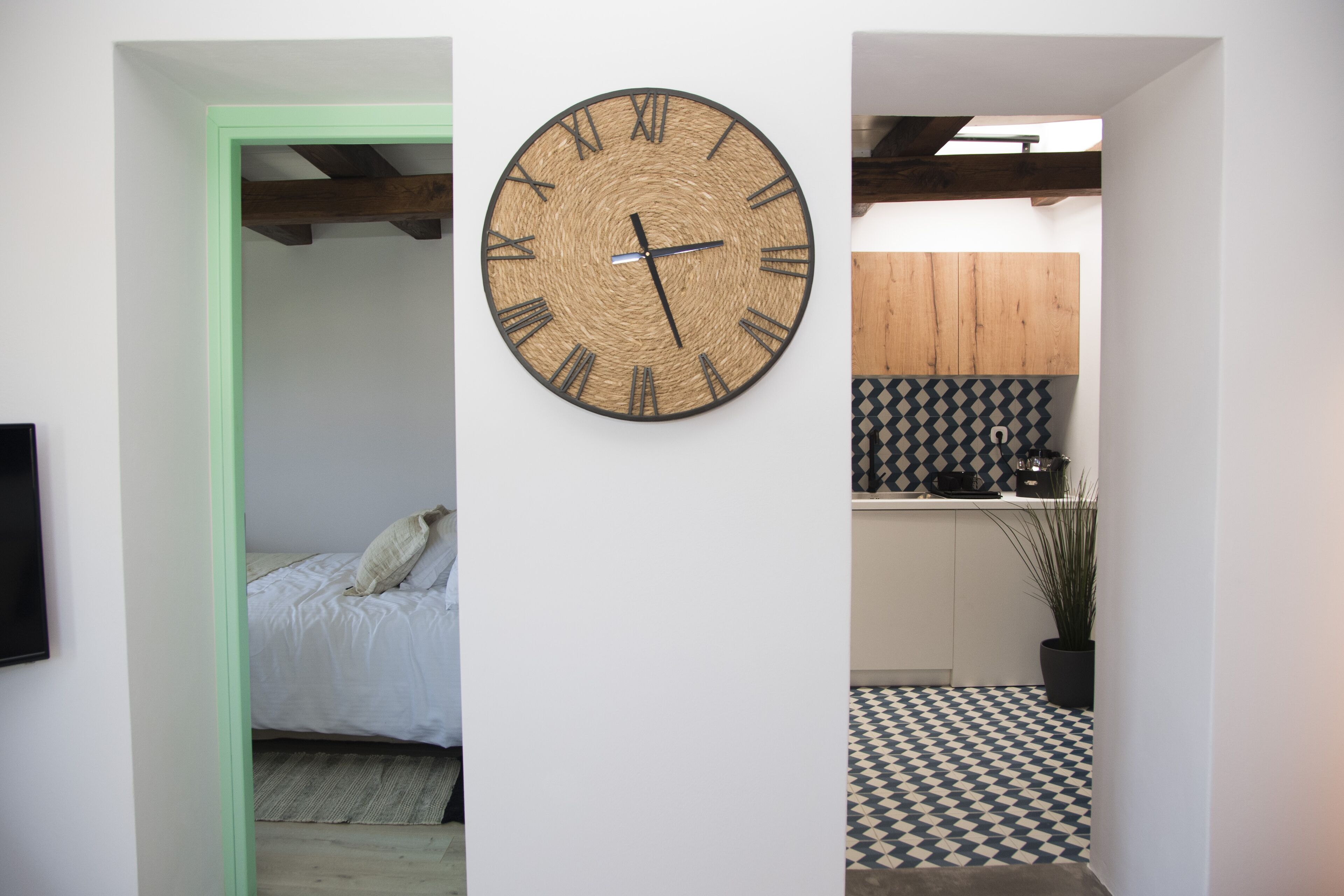 A room with a bed, a wall-mounted clock, and a kitchen area with a checkered floor.