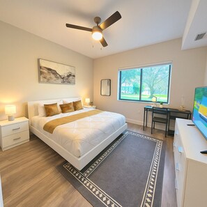 Een slaapkamer met een bed, een bureau, een stoel, een plafondventilator, een raam en een schilderij aan de muur.