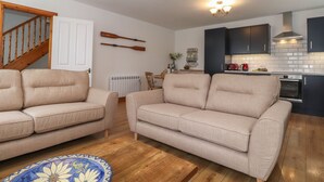 A modern living room with two beige sofas, a wooden coffee table, and a patterned rug. A kitchen with dark cabinets and a stove is visible in the background. - Coastal Nest (Seahouses)
