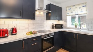 A modern kitchen with dark cabinets, a microwave, a toaster, and a window with nautical decor. - Coastal Nest (Seahouses)