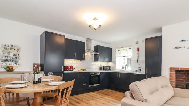 A modern kitchen with dark cabinets, a wooden dining table, and a beige sofa. - Coastal Nest (Seahouses)
