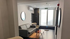 Un hall d’hôtel moderne avec un canapé gris, un miroir rond, une table basse en bois et une vue sur l’extérieur à travers une porte vitrée.