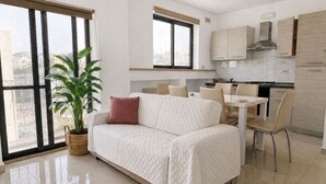 A modern living room with a white sofa, a dining table with chairs, a potted plant, and a kitchen area with cabinets and a stove. - Stylish 2BR Meridian suite in central St Julians (San Ġiljan)