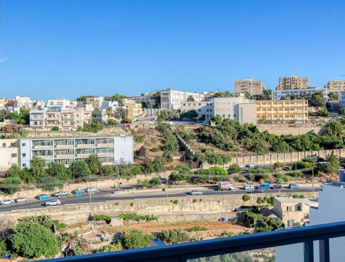 A coastal cityscape with modern buildings, a road, and greenery.