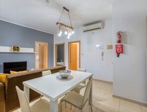 A modern living room with a sofa, dining table, and chairs. There is a television, a fireplace, and a ceiling light fixture. - Stylish 2BR Meridian suite in central St Julians (San Ġiljan)