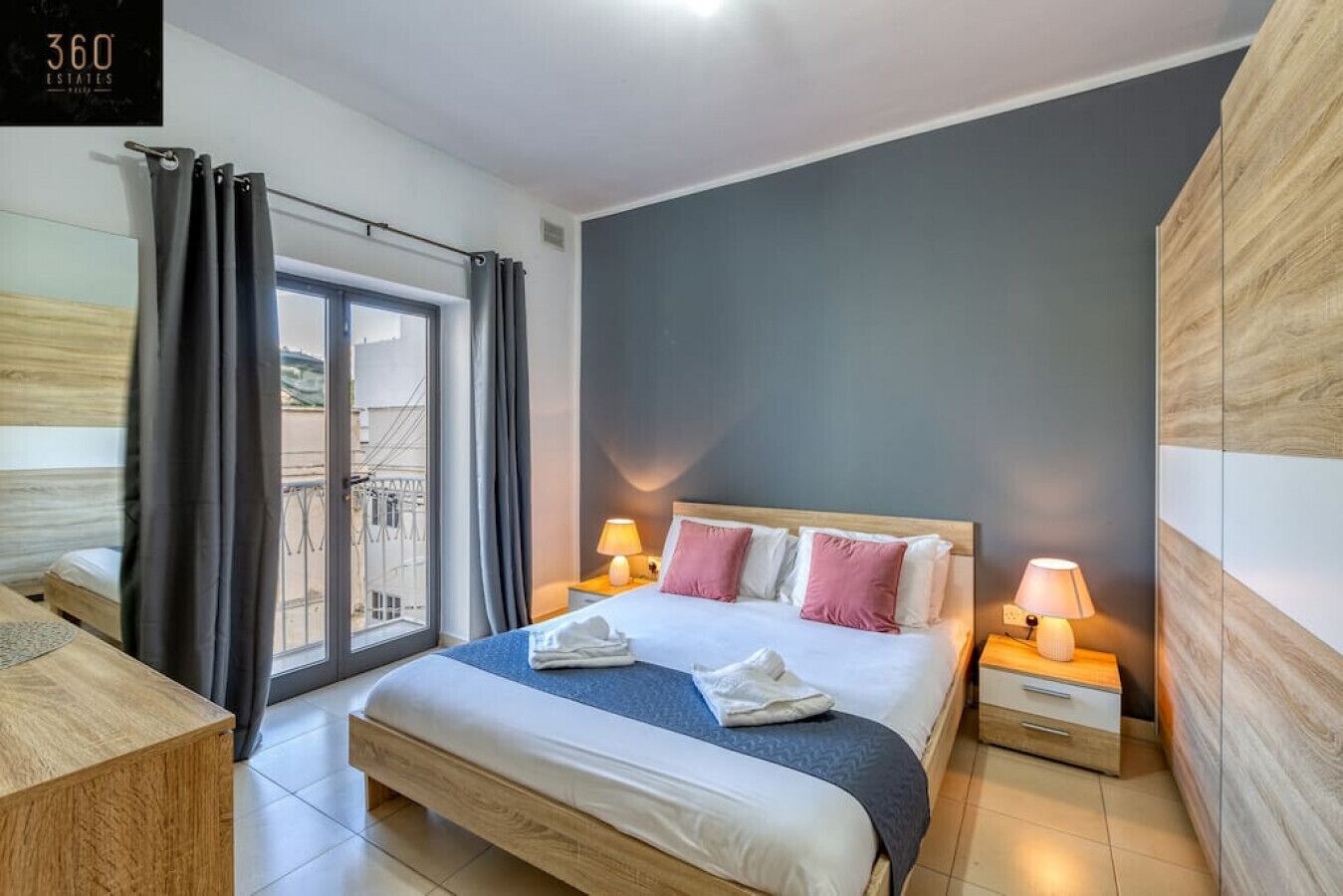 A modern hotel room with a large bed, two bedside tables with lamps, a view of the outside balcony, and a wooden headboard.