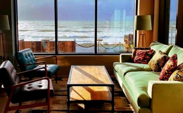 A room with a large window overlooking the ocean, a green sofa with colorful pillows, a wooden coffee table, and a blue armchair.
