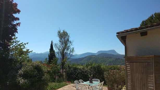 Eine Terrasse mit Tisch und Stühlen, umgeben von Grünpflanzen und Blick auf ferne Berge.