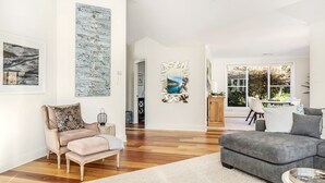 A spacious living room with hardwood flooring, a grey sofa, a beige armchair, and a coffee table. There are two large abstract paintings on the walls and a window with a view of greenery outside. - Hollydale, Burradoo, Southern Highlands (Burradoo)