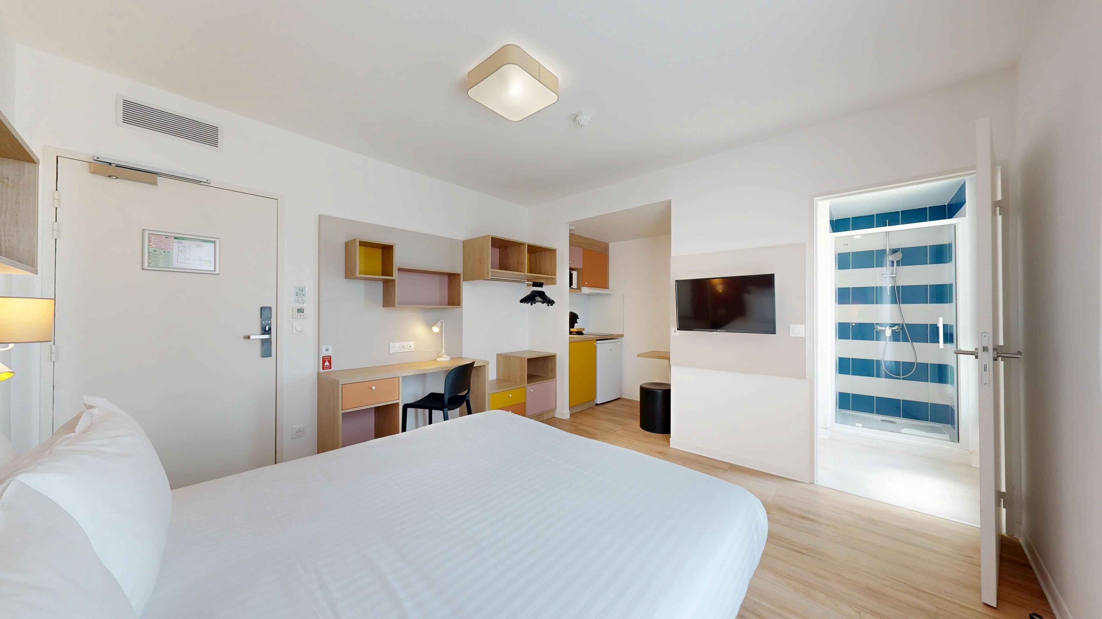Quarto de hotel moderno com uma cama grande, uma mesa com cadeira, TV fixada na parede e banheiro com chuveiro e porta de vidro.