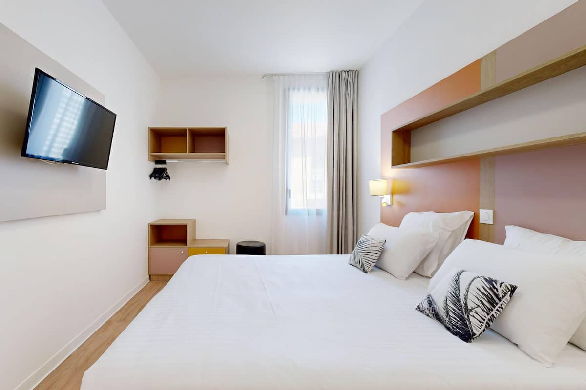 Une chambre d’hôtel avec un grand lit, un téléviseur à écran plat fixé au mur, une table de chevet avec une lampe et une fenêtre avec des rideaux.