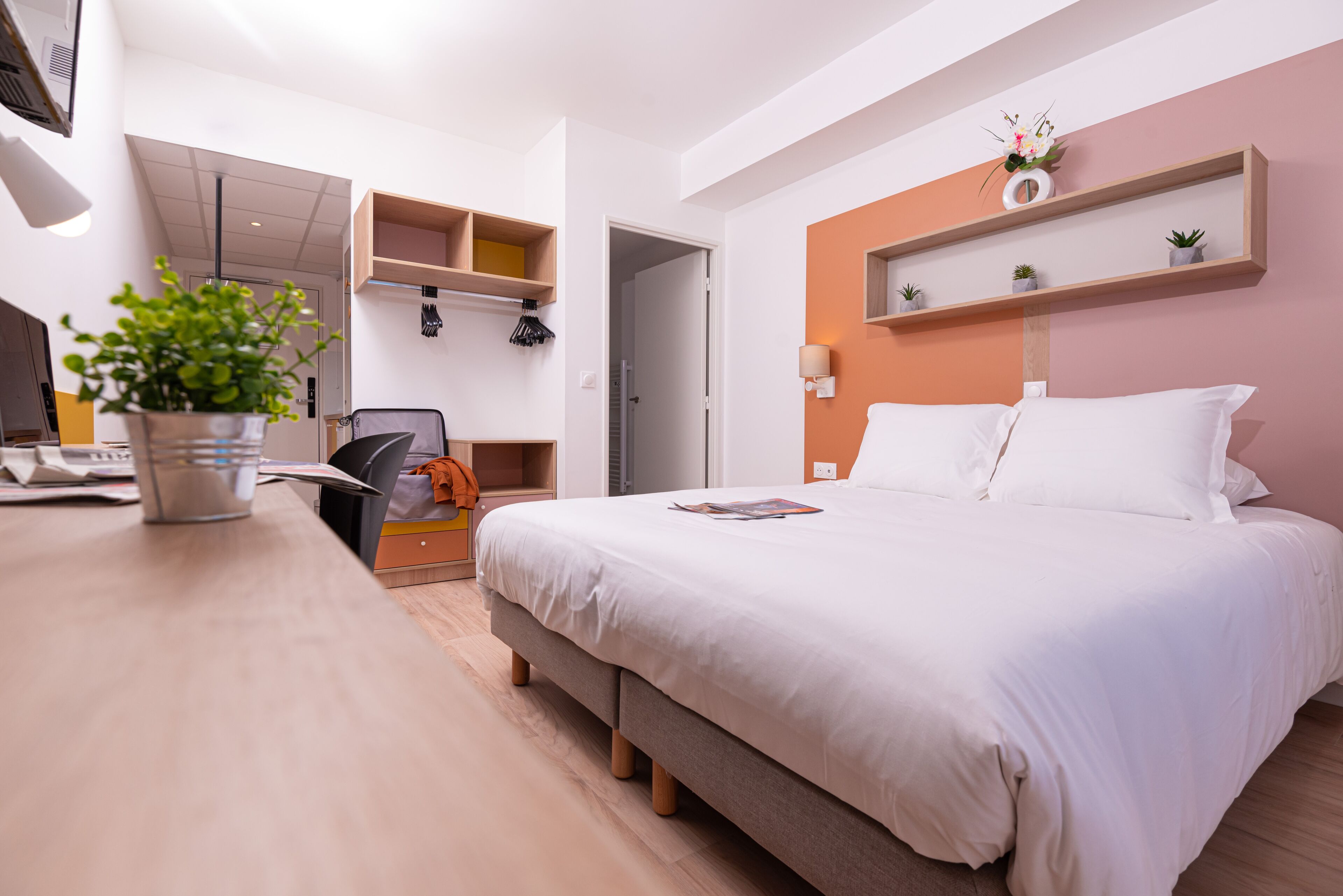 A modern hotel room with a bed, a desk with a computer, a chair, and a potted plant.