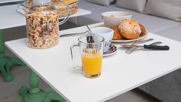 A breakfast table with a glass pitcher, a glass of orange juice, a bowl of cereal, a croissant, and a loaf of bread.