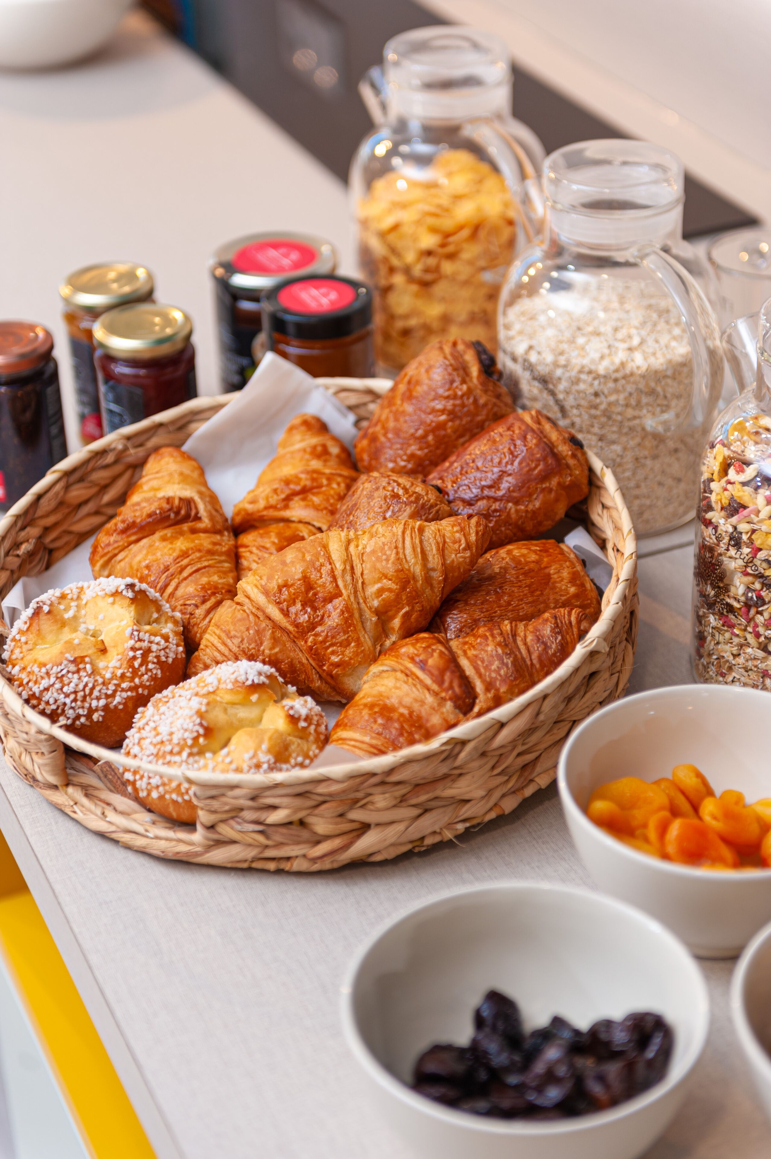 A basket of croissants and pastries, jars of jams, and bowls of fruits on a table.