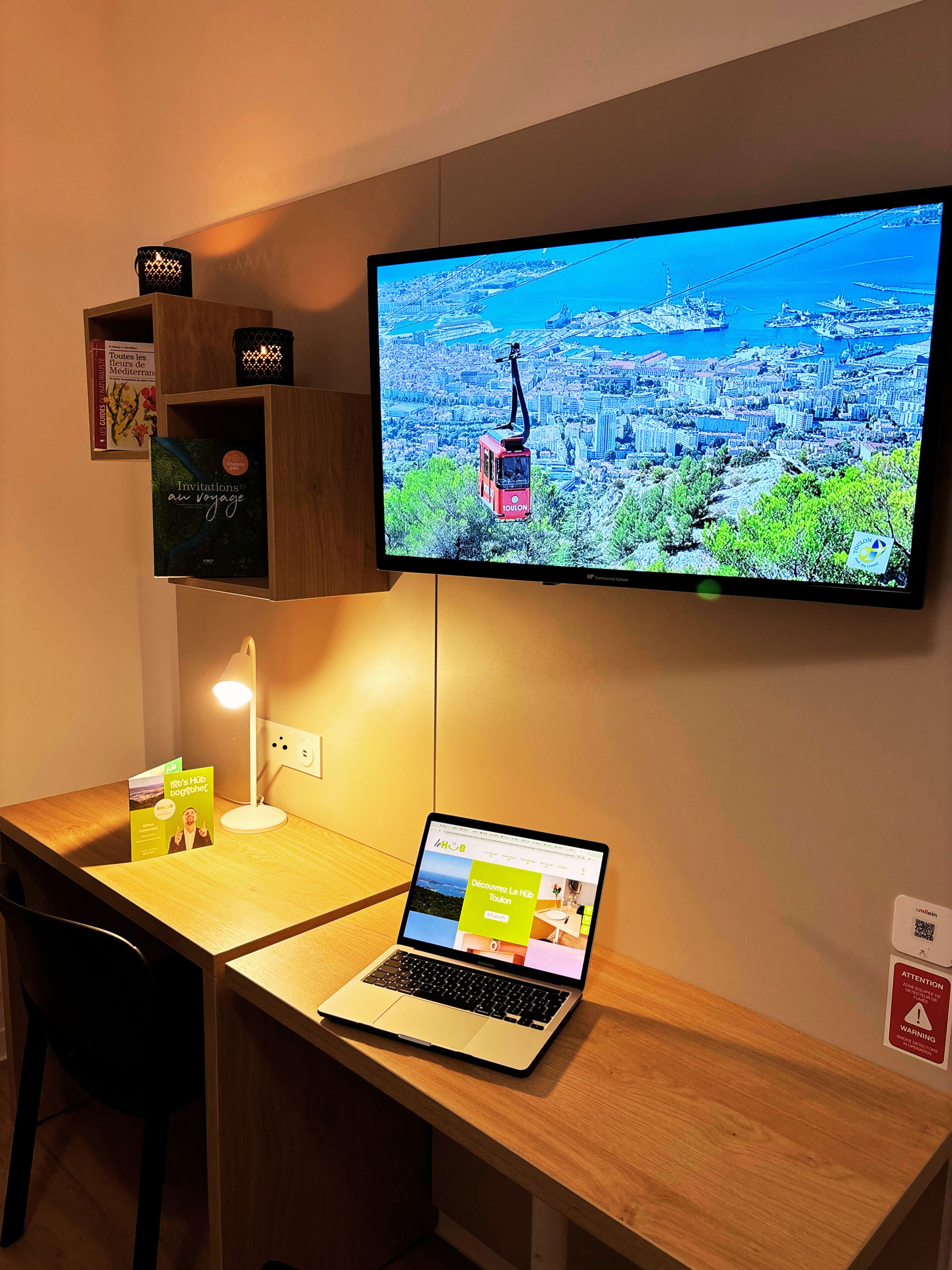 A hotel room with a wooden desk, a laptop, a television displaying a cityscape, a bookshelf with books, and a small table lamp.