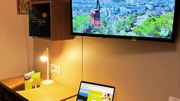 A hotel room with a wooden desk, a laptop, a television displaying a cityscape, a bookshelf with books, and a small table lamp.