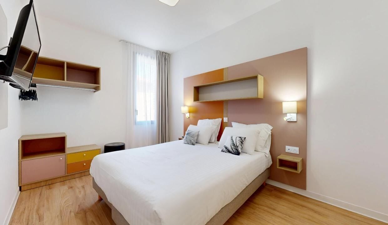 A modern hotel room with a large bed, bedside tables, a desk, and a wardrobe.