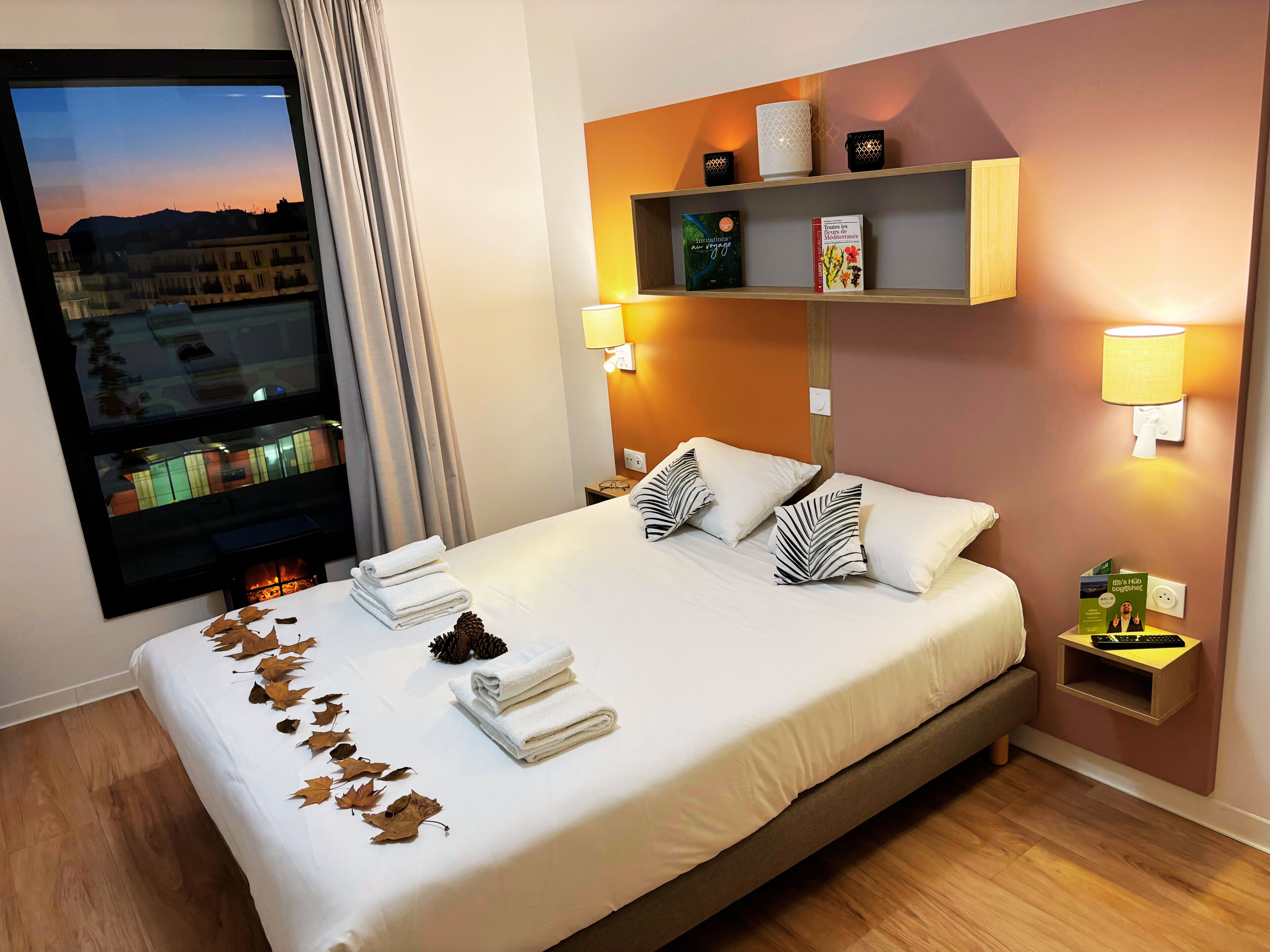 A neatly made bed with white linens, a nightstand with a lamp, and a shelf with books and decorative items.