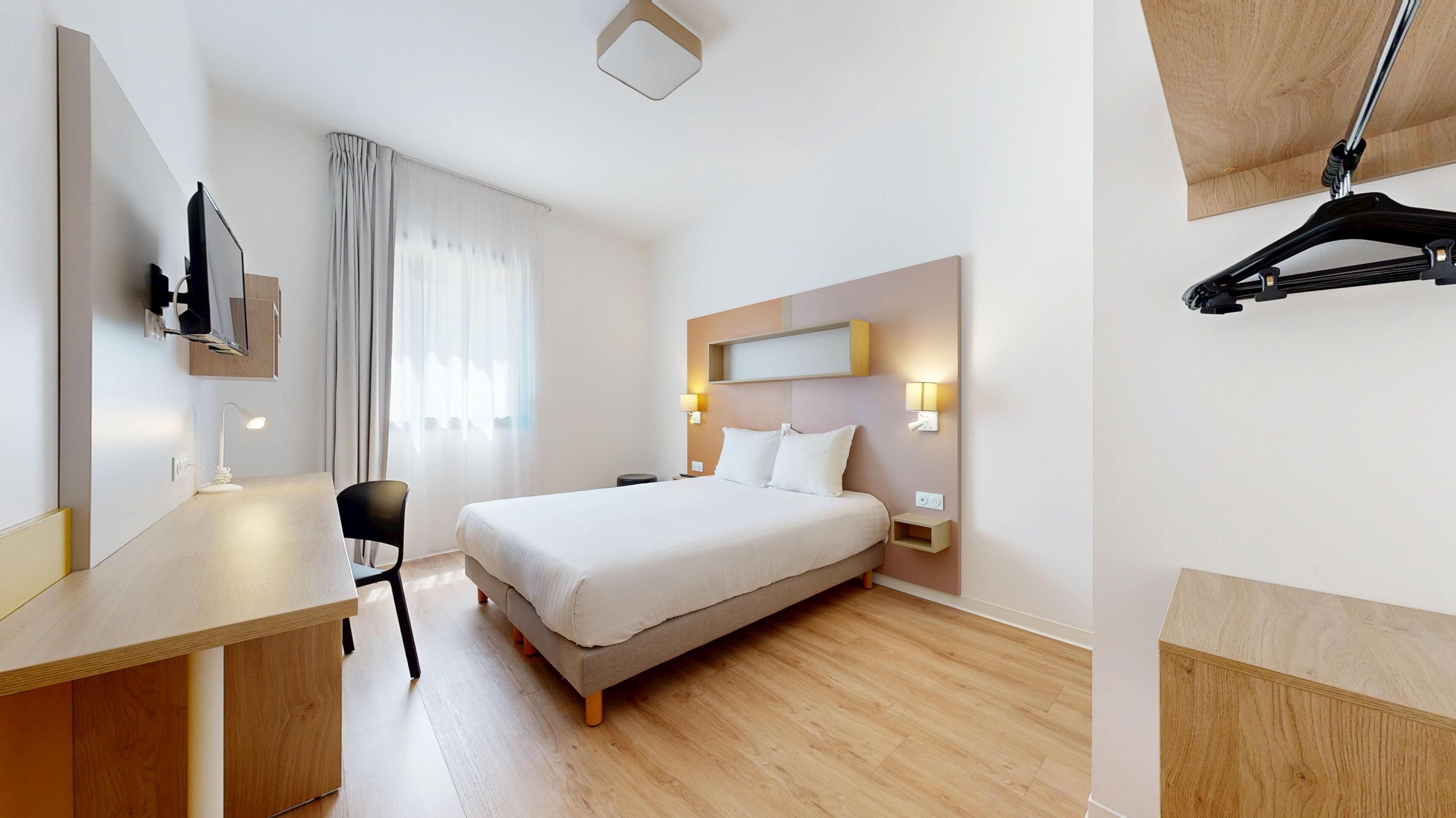 Ett hotellrum med en säng, ett skrivbord, en stol och en tv som är monterad på väggen.