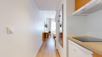 A narrow hallway with a wooden floor, a desk with a chair, a bed, and a white wall with a light switch.