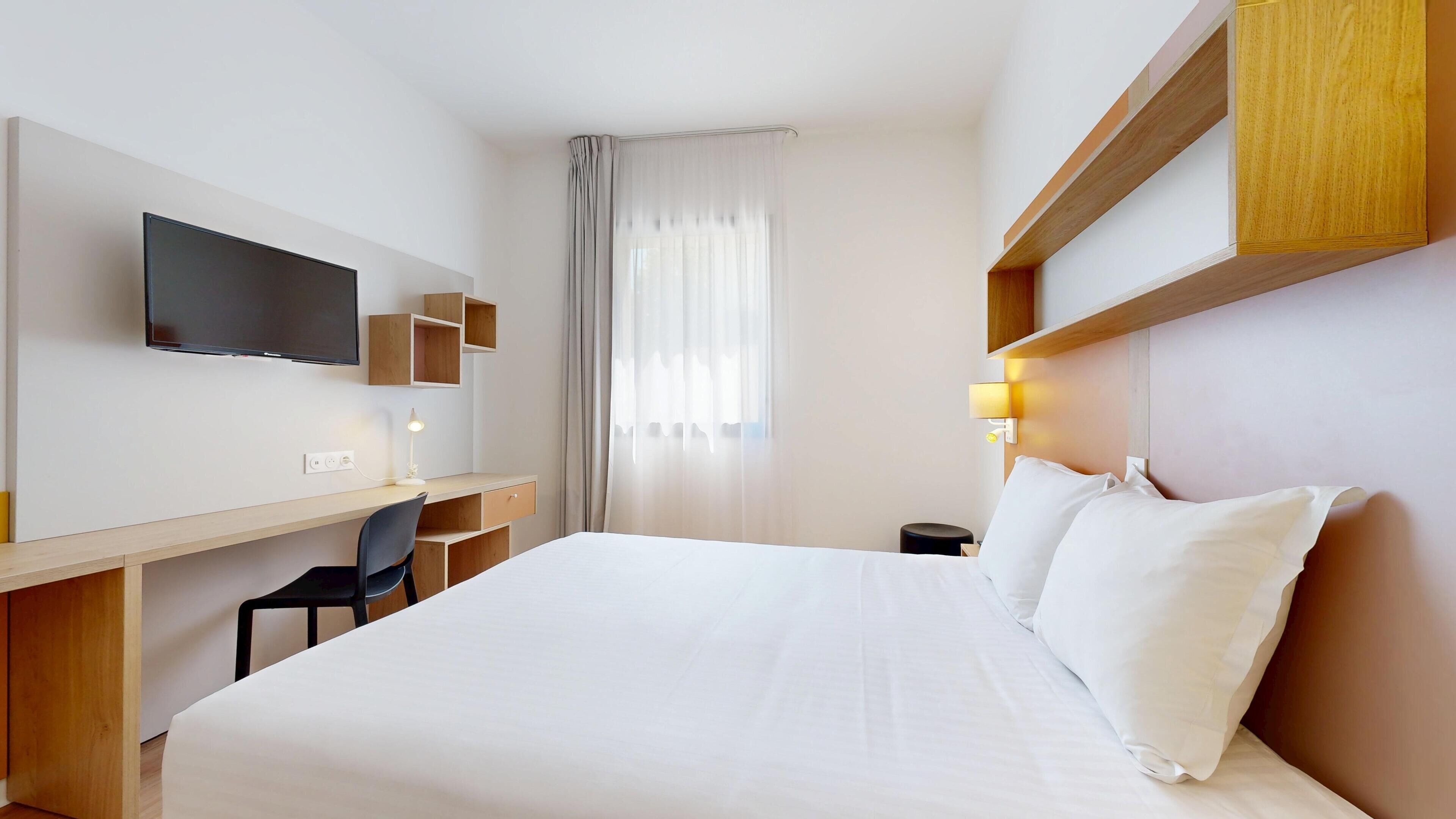 A hotel room with a bed, a desk with a chair, a television, and a window with curtains.