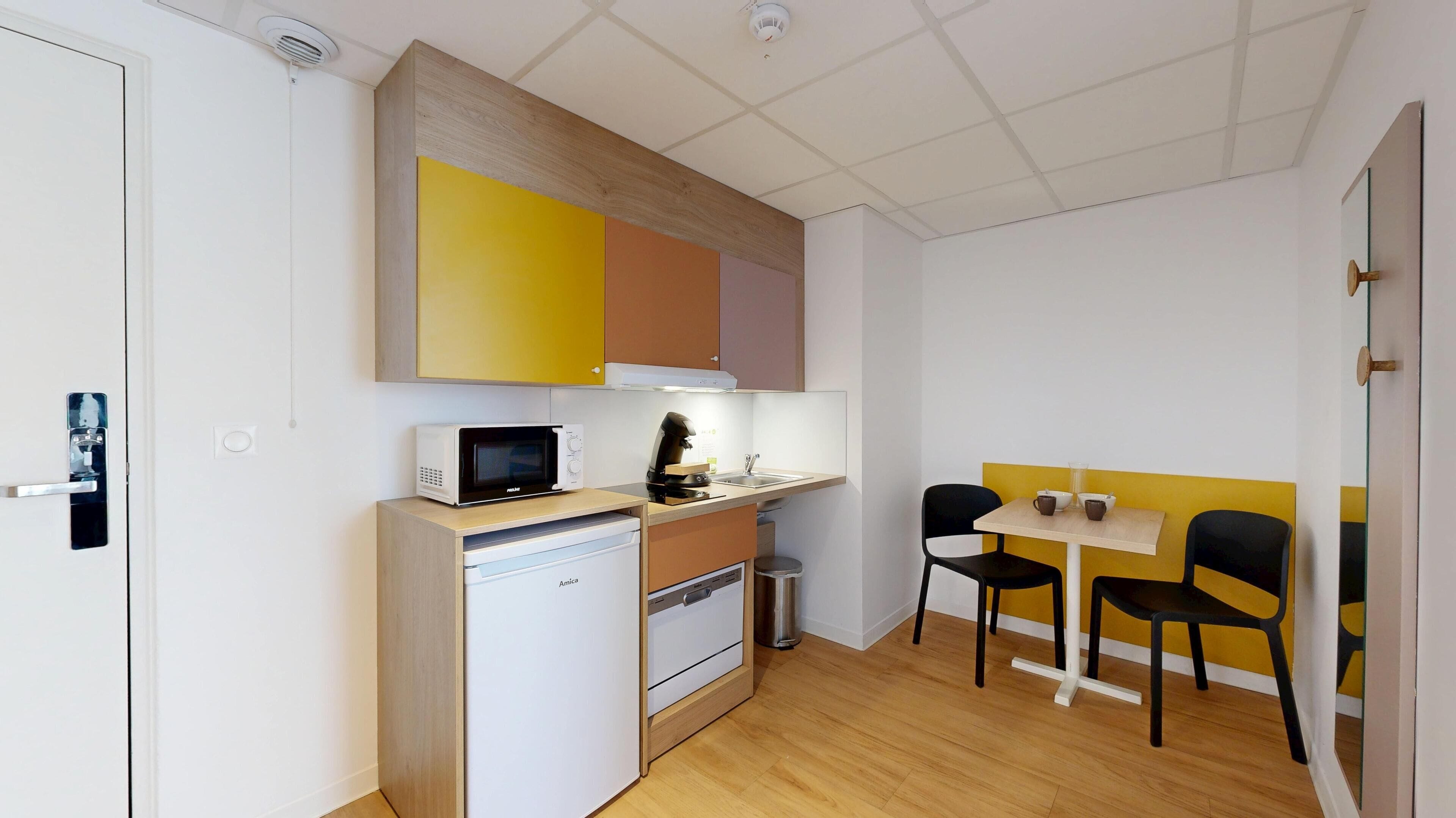 Cocina compacta con microondas, cafetera y un lavamanos pequeño. Hay una zona de comedor con dos sillas y una mesa.