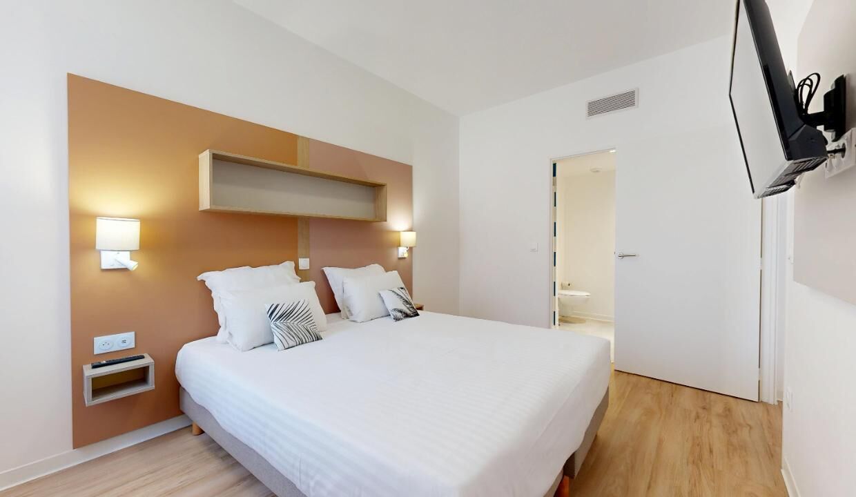 A modern hotel room with a large bed, bedside lamps, a built-in shelf, and a flat-screen TV mounted on the wall.