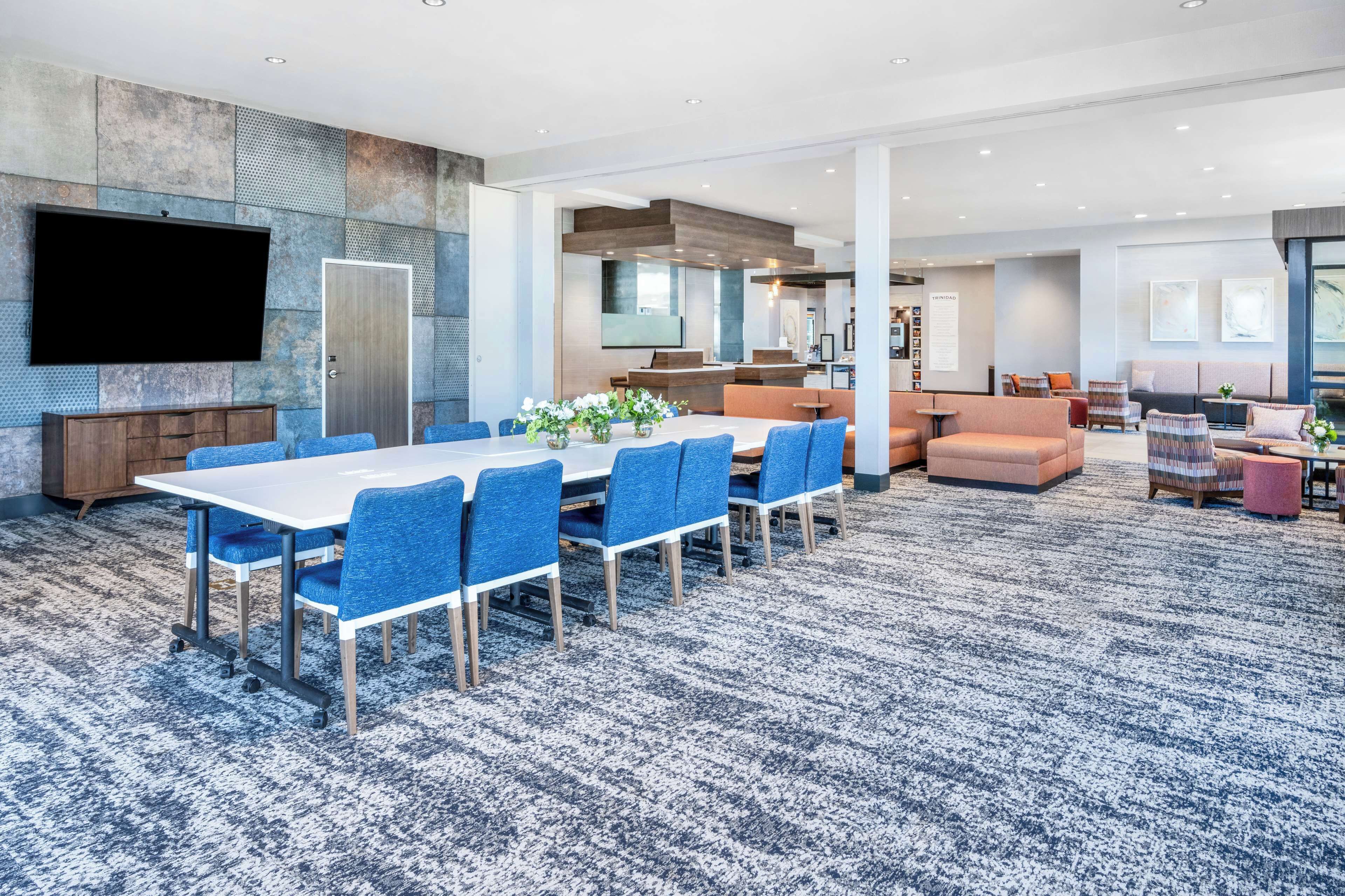 A spacious hotel lobby with a large conference table, blue chairs, a flat-screen TV, and a modern kitchen area in the background.