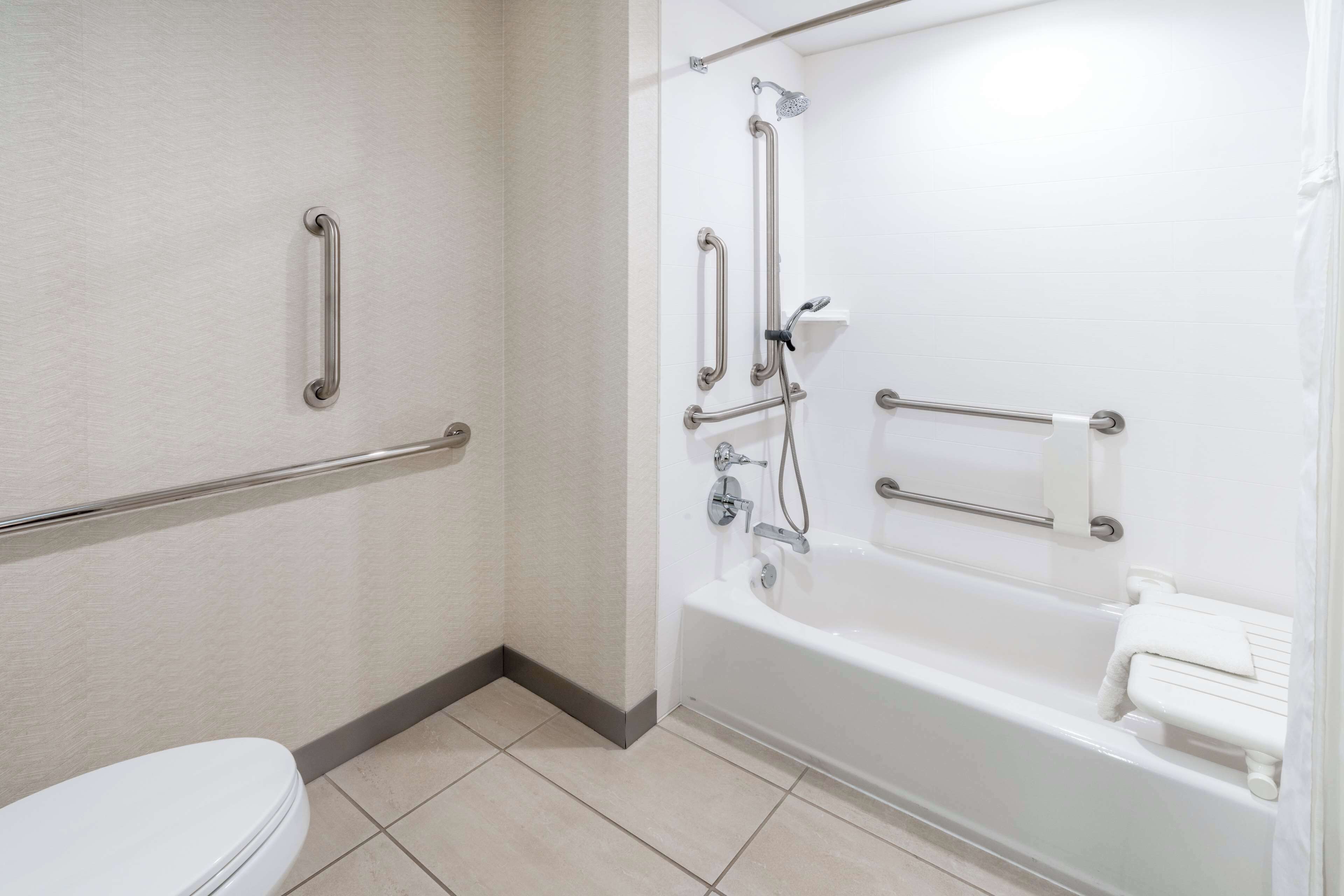 Une salle de bain avec des toilettes blanches, une baignoire équipée d’un pommeau de douche et des barres d’appui fixées aux murs.