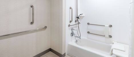 Une salle de bain avec des toilettes blanches, une baignoire équipée d’un pommeau de douche et des barres d’appui fixées aux murs.