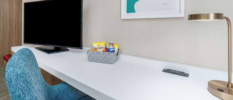 A hotel room with a flat-screen TV, a framed abstract art piece, a desk with a lamp, and a basket of snacks.