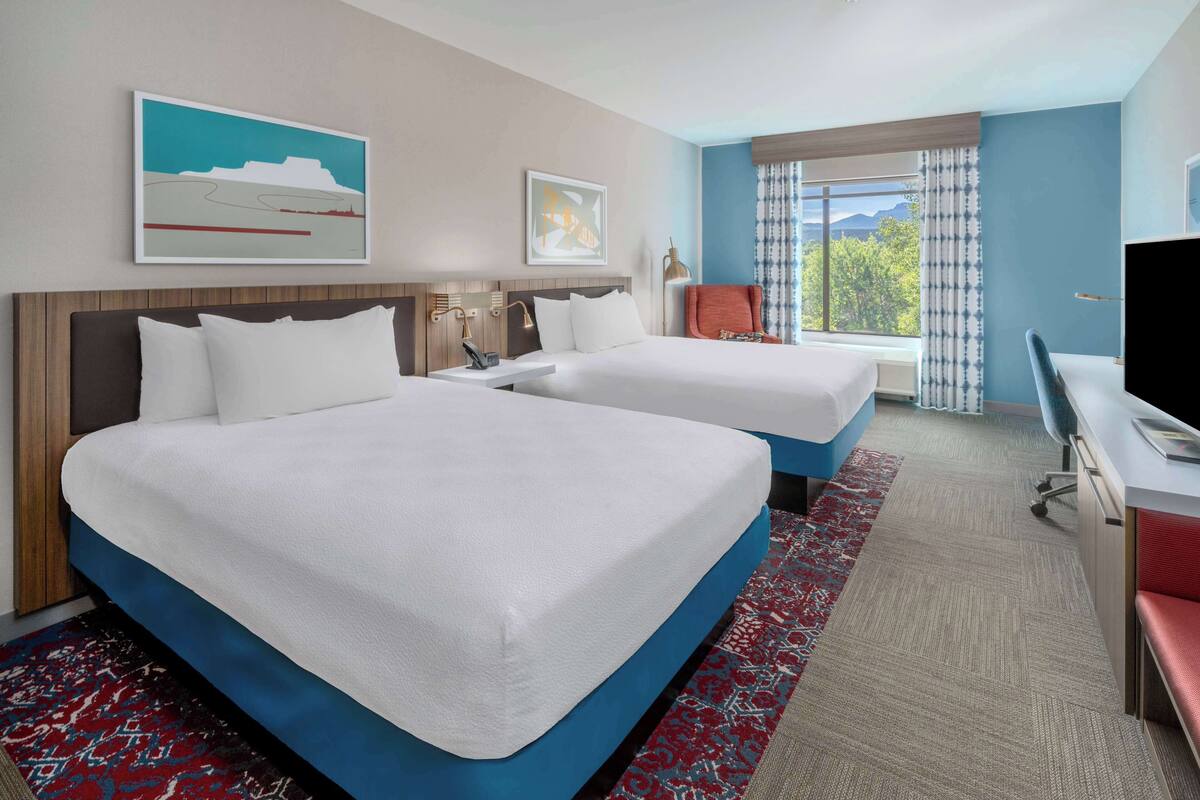 A hotel room with two beds, a desk, a chair, and a TV.