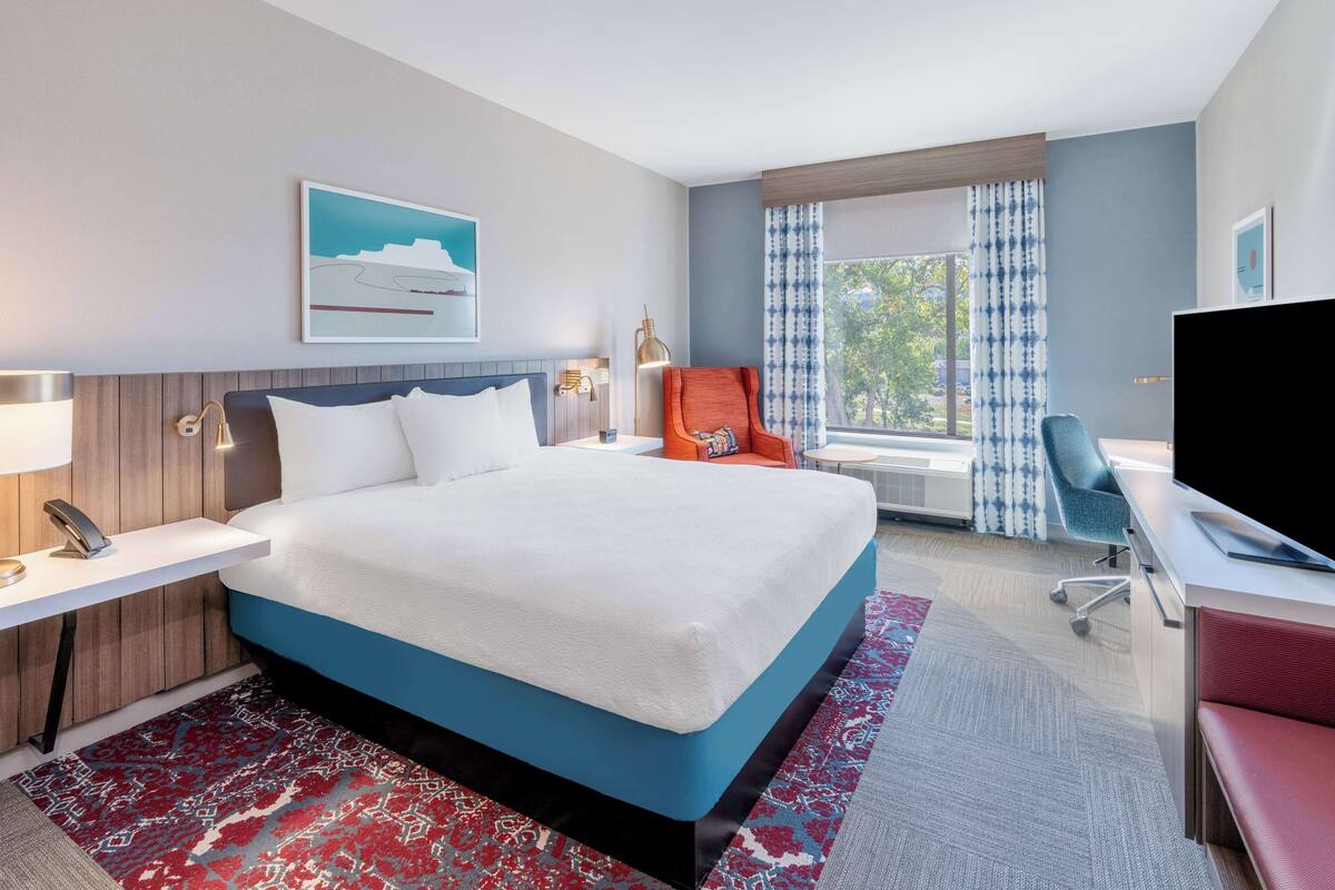 A hotel room with a large bed, a desk with a computer, a TV, and a window with curtains.
