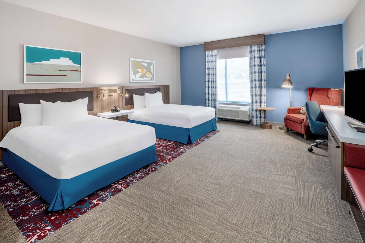 A hotel room with two beds, a desk, a chair, a TV, and artwork on the walls.